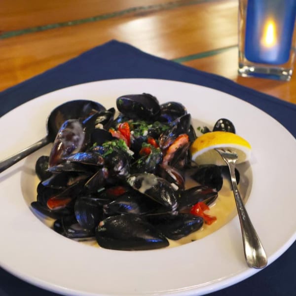 A white plate of mussels in a creamy sauce with red peppers, parsley, and a lemon wedge.