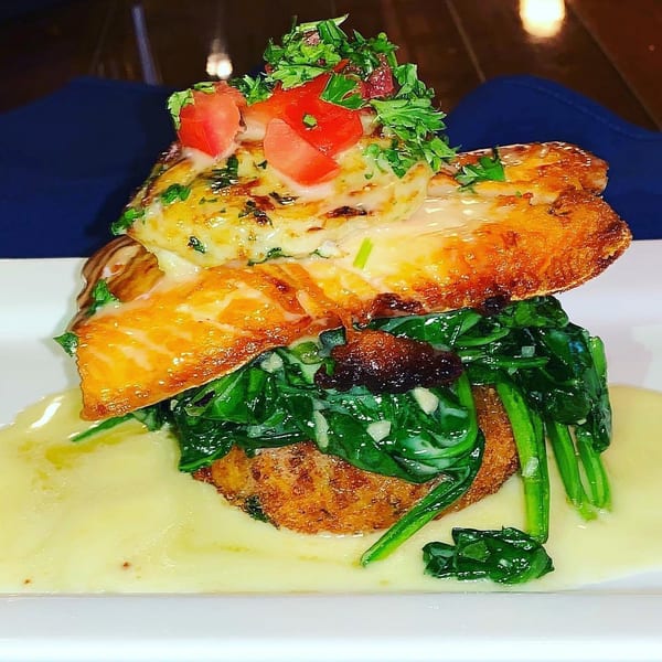 Pan-seared salmon with melted cheese, diced tomatoes, and parsley, atop spinach and a fried patty, with a creamy sauce.